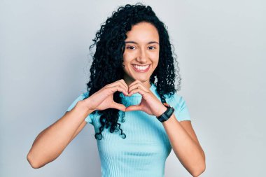 Kıvırcık saçlı, sıradan mavi tişörtlü, aşık gülümseyen, elleriyle kalp sembolü yapan genç İspanyol kadın. romantik kavram. 