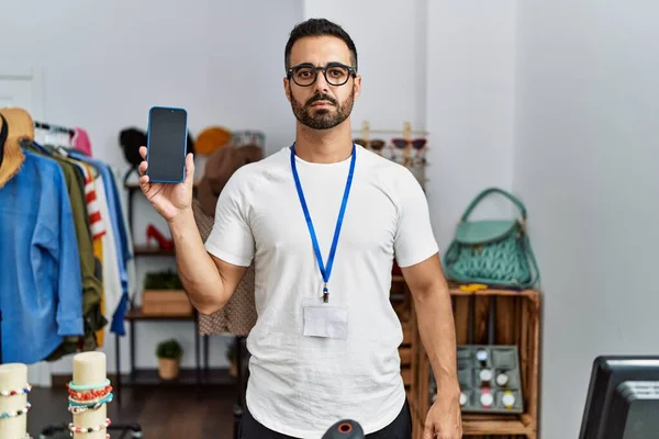 小売店の店長を務める髭を生やした若いヒスパニック系の男がスマホ思考の姿勢と落ち着いた表情で自信を持っている — ストック写真