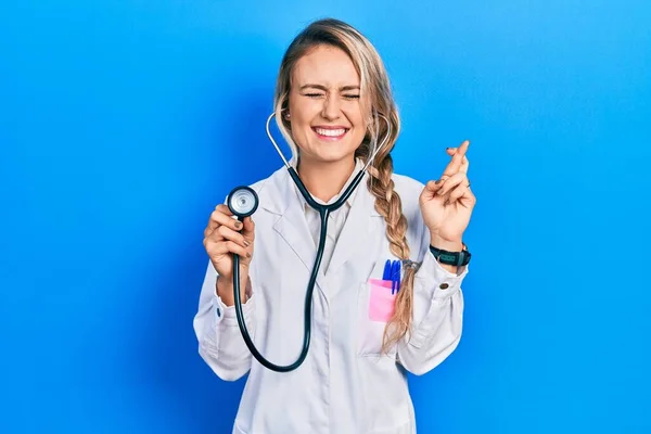 Beautiful Young Blonde Doctor Woman Holding Stethoscope Gesturing Finger Crossed — Stock Photo, Image