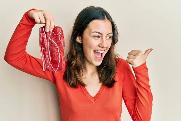 Joven Morena Sosteniendo Carne Cruda Señalando Pulgar Hacia Lado Sonriendo —  Fotos de Stock