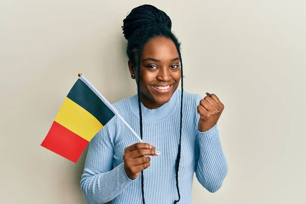African american woman with braided hair holding belgium flag screaming proud, celebrating victory and success very excited with raised arm