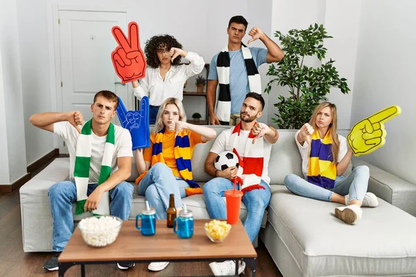 Group of friends supporting football team watching tv al home sitting on the sofa with angry face, negative sign showing dislike with thumbs down, rejection concept