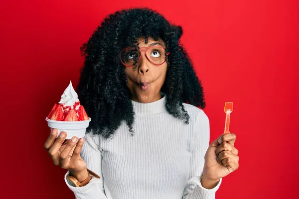 African american woman with afro hair eating strawberry ice cream making fish face with mouth and squinting eyes, crazy and comical.