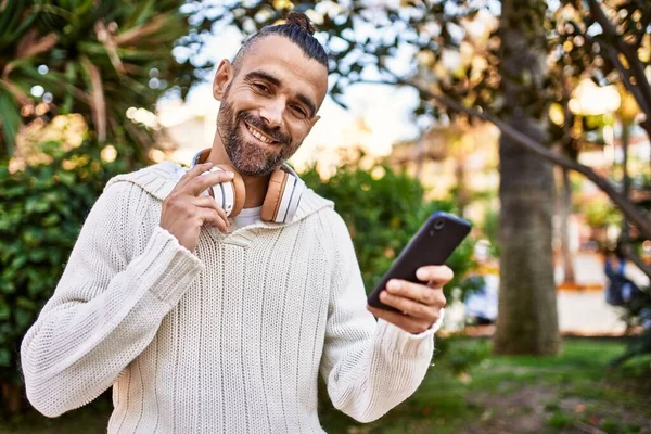 髭を生やしたハンサムな中年男性は スマートフォンやヘッドフォンを使用して幸せと自信を持って屋外に立っています — ストック写真
