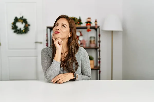 Young latin woman sitting on the table by christmas decor looking confident at the camera smiling with crossed arms and hand raised on chin. thinking positive.