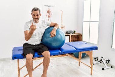 Orta yaşlı İspanyol bir adam ağrı kesici kliniğinde elinde pilates topu tutarak el kol hareketi yaparak mutlu bir şekilde karşılamaya ve gülümsemeye davet ediyor. 