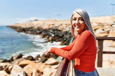 Orta yaşlı, kır saçlı, kumsalda mutlu gülümseyen bir kadın..