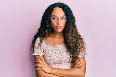 Gündelik kıyafetler giyen, gözlüklü, kuşkucu ve gergin genç Latin bir kadın. Yüzünde çapraz kollarla onaylamayan bir ifade var. negatif kişi. 