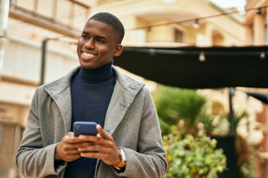 Genç Afrikalı Amerikalı adam şehirde akıllı telefon kullanarak mutlu bir şekilde gülümsüyor.