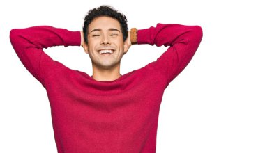 Rahat ve esnek giyinen genç yakışıklı adam, kolları ve elleri başının arkasında mutlu bir şekilde gülümsüyor. 