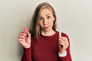 Genç sarışın kadın, elinde regl fincanı ve tampon tutarken şüpheci ve gergin, sorun yüzünden somurtuyor. negatif kişi. 