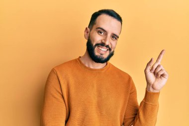 Sakallı, sıradan bir kazak giyen, yüzünde kocaman bir gülümseme olan, eli ve parmağı kameraya bakan genç bir adam.. 