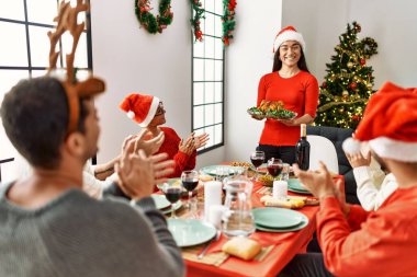Bir grup insan el çırpıyor ve masada oturuyor. Ayakta duran ve elinde kavrulmuş hindi tutan kadın Noel 'i evde kutluyor..
