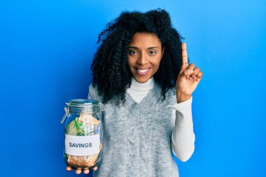 Afro saçlı Afrikalı bir kadın, elinde tasarruf kavanozuyla Güney Afrika 'dan para getiriyor. Bir fikir veya soruyla gülümsüyor ve mutlu bir yüzle işaret ediyor. 
