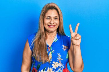 Orta yaşlı İspanyol kadın gündelik kıyafetler giyip kameraya gülümseyen ve zafer işareti yapan. İki numara.. 