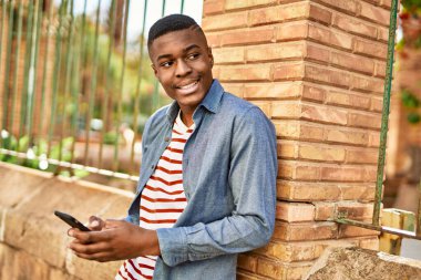 Genç Afrikalı Amerikalı adam şehirde akıllı telefon kullanarak mutlu bir şekilde gülümsüyor..