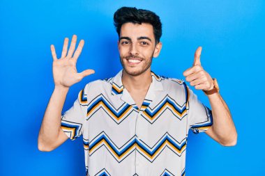 Latin asıllı genç adam gündelik giysiler giyip altı numaralı parmaklarıyla işaret ederken kendinden emin ve mutlu bir şekilde gülümsüyor.. 
