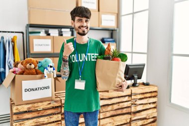 Bağış noktasında gönüllü tişört giyen İspanyol sakallı adam mutlu ve olumlu gülümsüyor, başparmağını kaldırıp mükemmel ve onay işareti yapıyor. 