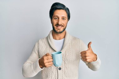 Genç İspanyol adam bir fincan kahve içiyor mutlu ve pozitif gülümsüyor, başparmağıyla mükemmel ve onay işareti yapıyor. 