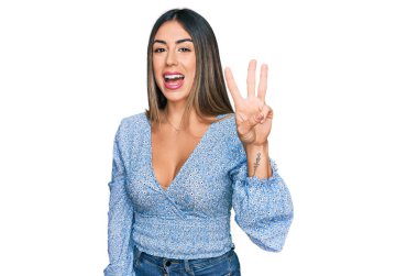 Genç İspanyol kadın gündelik kıyafetler giyer ve üç numaralı parmaklarıyla gülümseyerek mutlu ve kendinden emin bir şekilde işaret eder.. 