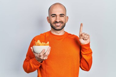 Elinde patates cipsi olan genç kel bir adam bir fikirle gülümsüyor ya da bir soru işareti yapıyor. 