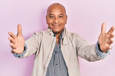 Orta yaşlı Latin bir adam, sıradan kıyafetler giyip kameraya bakıyor ve sarılmak için kollarını açmış gülümsüyor. Mutluluğu kucaklayan neşeli bir ifade.. 