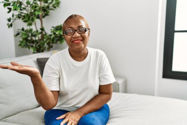 Olgun İspanyol kadın evdeki kanepede oturuyor neşeli bir şekilde gülümsüyor ve avuç içi kameraya bakıyor.. 