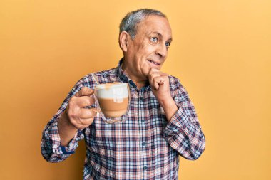 Gri saçlı, eli çenesinde fincan kahve içen, soru sormayı düşünen yakışıklı bir son sınıf öğrencisi. Düşünceli bir ifade. Gülümseyen ve düşünceli bir yüz. şüphe kavramı. 