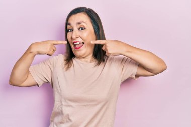 Orta yaşlı İspanyol kadın gündelik kıyafetler giyip neşeli bir şekilde gülümsüyor ve parmaklarıyla ve ağzıyla işaret ediyor. diş sağlığı kavramı. 