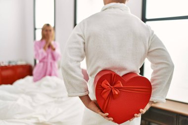 Sevgilisine sürpriz yapan bir adam sevgilisinin hediye kutusuyla evde yatağında oturuyor..