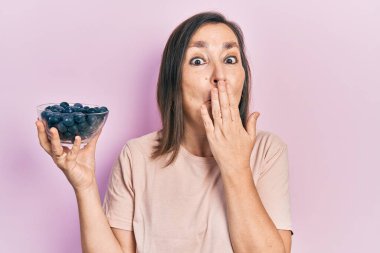 Orta yaşlı İspanyol bir kadın elinde yaban mersini tutarak ağzını eliyle kapatıyor, şokta ve hata yapmaktan korkuyor. şaşırmış ifade 