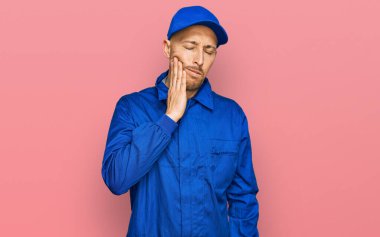 Sakallı, inşaatçı tulumu giyen kel bir adam diş ağrısı ya da diş hastalığından dolayı ağızlarına acı dolu bir ifadeyle dokunuyor. dişçi 