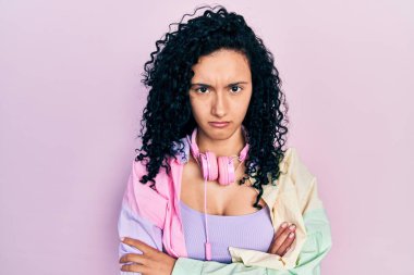 Kıvırcık saçlı, spor kıyafetleri giyen, kulaklık takan kuşkucu ve gergin, yüzünde çapraz kollu tasvip etmeyen İspanyol bir kadın. negatif kişi. 