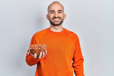 Fıstık tutan genç kel adam pozitif ve mutlu görünüyor. Duruyor ve gülümsüyor. Kendine güvenen bir gülümsemeyle dişlerini gösteriyor. 