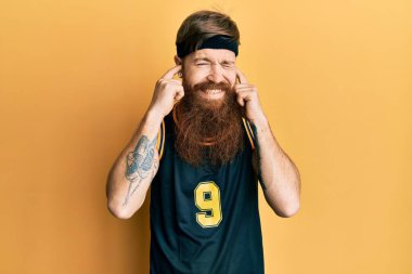 Uzun sakallı, basketbol forması giyen, kulaklarını parmaklarıyla kapatan, yüksek sesli müzik sesi için rahatsız edici ifadeleri olan kızıl saçlı bir adam. sağır kavramı. 