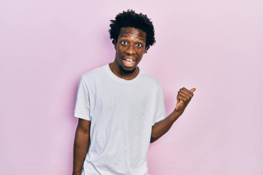 Beyaz bir tişört giyen, gülümseyen, gülümseyen ve baş parmağıyla yüzünü gösteren Afro-Amerikan genç bir adam.. 