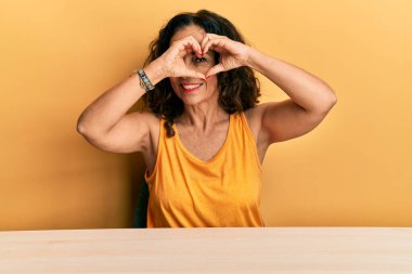 Güzel orta yaşlı kadın, gündelik giysiler içinde masada oturuyor kalp şeklinde oturuyor, elleri ve parmaklarıyla gülümsüyor ve tabelanın içinden bakıyor. 