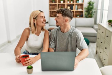 Genç beyaz çift dizüstü bilgisayar kullanıyor ve kahve içiyor. Evdeki masada oturuyorlar..