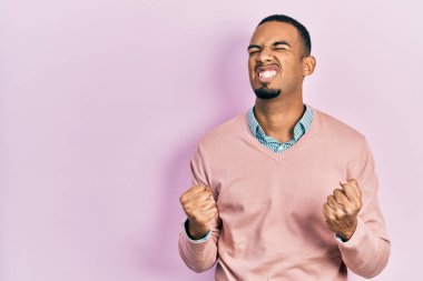 Zarif kıyafetler giyen, çok mutlu ve heyecanlı genç bir Afro-Amerikan erkeği kolları havada, gülümseyen ve başarı için çığlık atan, kazanan bir jest yapıyor. kutlama konsepti. 