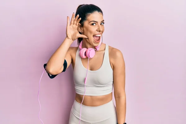 若いヒスパニック系の女性はジムの服を着て 噂やゴシップに耳を傾ける耳の上に手で笑みを浮かべてヘッドフォンを使用しています 聴覚障害の概念 — ストック写真