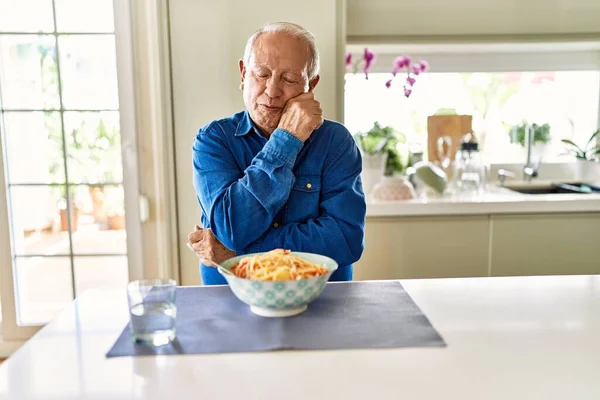 Senior man with grey hair eating pasta spaghetti at home thinking looking tired and bored with depression problems with crossed arms.