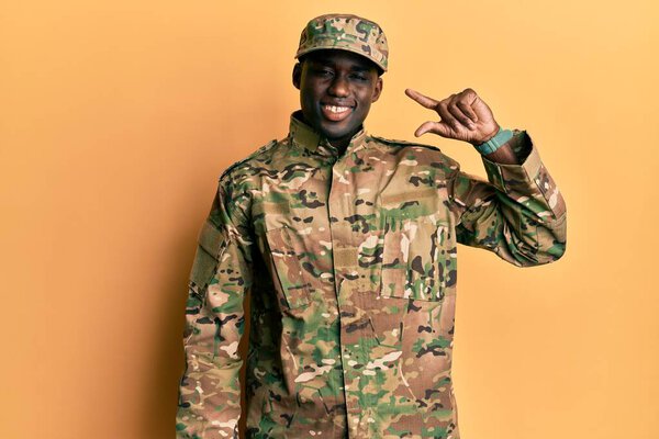 Young african american man wearing army uniform smiling and confident gesturing with hand doing small size sign with fingers looking and the camera. measure concept. 