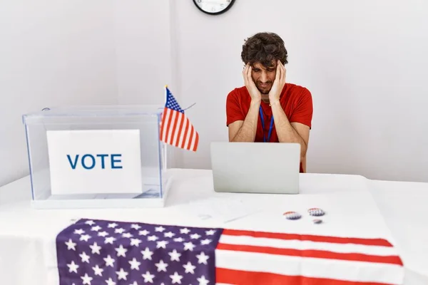 Young hispanic man at political election sitting by ballot with hand on head for pain in head because stress. suffering migraine.