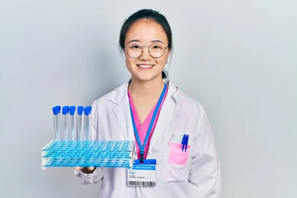 Young chinese girl wearing scientist uniform holding test tube looking positive and happy standing and smiling with a confident smile showing teeth