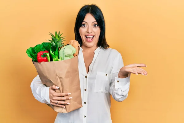 年轻的惊慌失措的女人拿着装有食品杂货的纸袋 开心地微笑着庆祝胜利 高举双手庆祝胜利 — 图库照片