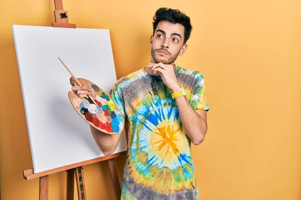 Young hispanic man standing drawing with palette by painter easel stand with hand on chin thinking about question, pensive expression. smiling with thoughtful face. doubt concept.