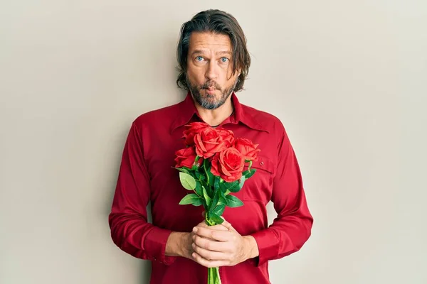 Middle age handsome man holding bouquet of red roses making fish face with mouth and squinting eyes, crazy and comical.