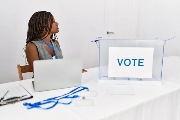 Young african american woman working at political election sitting by ballot looking to side, relax profile pose with natural face and confident smile.