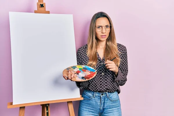 画家のイーゼルによるパレットで絵を描く美しいヒスパニック系の女性は 無知で混乱した表情を立っています 疑わしい概念 — ストック写真