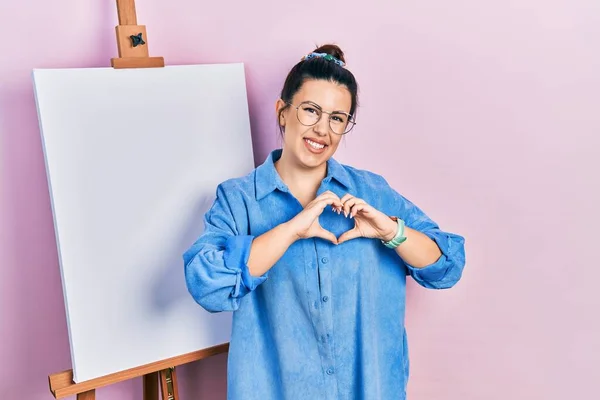 画家のイーゼルのそばに立つ若いヒスパニック系の女性は手で心のシンボル形状を行う愛に笑みを浮かべて立っている ロマンチックなコンセプト — ストック写真